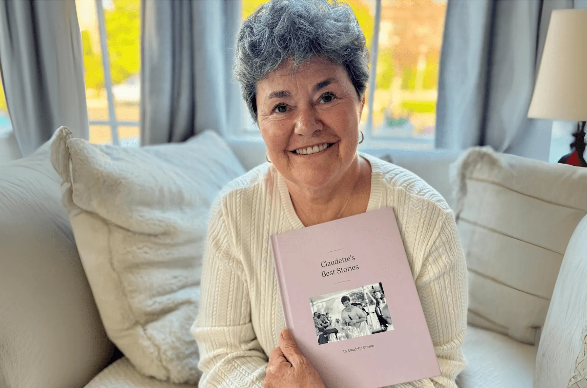 An older woman with short gray hair, sitting comfortably on a white couch, smiling warmly while holding a pink book titled 'Claudette's Best Stories' with a black-and-white photo on the cover. The cozy living room features soft lighting and a large window in the background.